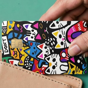 May include: A rectangular credit card featuring a vibrant, multi-colored doodle pattern of various cats in black, white, yellow, blue, red, and pink. The card includes a gold-colored EMV chip and the text DON'T printed vertically on the left side.
