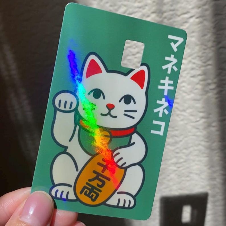 May include: A rectangular green card featuring a white Maneki-neko cat illustration with red ears, a red collar, and a yellow oval coin marked with black Japanese characters. The text "マネキネコ" is printed vertically in white on the right side.