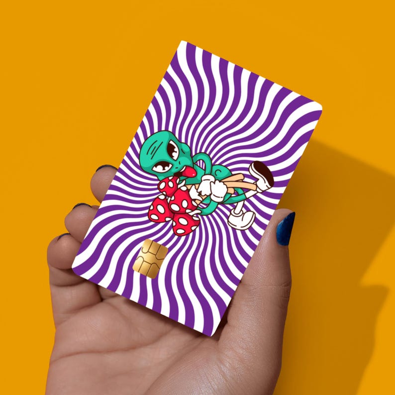 May include: A rectangular credit card skin featuring a vibrant purple and white wavy spiral pattern. In the centre, a green cartoon alien with large black eyes is depicted holding a cluster of red mushrooms with white spots, wearing white gloves.