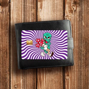 May include: A black bi-fold wallet featuring a credit card skin with a purple and white radial spiral background. A green cartoon alien with a red tongue is depicted holding a cluster of red and white spotted mushrooms in its arms.