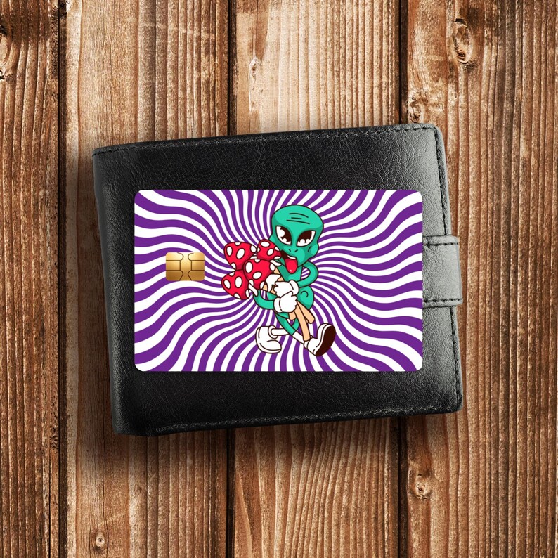 May include: A black bi-fold wallet featuring a credit card skin with a purple and white radial spiral background. A green cartoon alien with a red tongue is depicted holding a cluster of red and white spotted mushrooms in its arms.