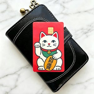 May include: A rectangular credit card skin featuring a white beckoning cat on a bright red background. The cat wears a green collar with a gold bell and holds a gold oval coin with black Chinese characters. A gold EMV chip is visible at the top.