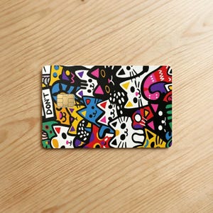 May include: A rectangular credit card skin featuring a vibrant, multi-colored doodle pattern of various cat faces in black, white, yellow, blue, red, and pink. The design includes a gold-colored chip and the text DON'T printed in white on black.