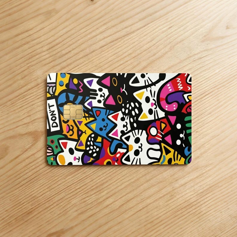 May include: A rectangular credit card skin featuring a vibrant, multi-colored doodle pattern of various cat faces in black, white, yellow, blue, red, and pink. The design includes a gold-colored chip and the text DON'T printed in white on black.