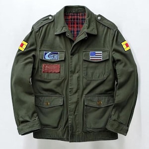 May include: An olive green field jacket featuring a red and blue plaid interior lining, shoulder epaulets, and four front pockets. The chest displays patches including an American flag and text reading Fast End. Yellow and red square patches adorn each sleeve.