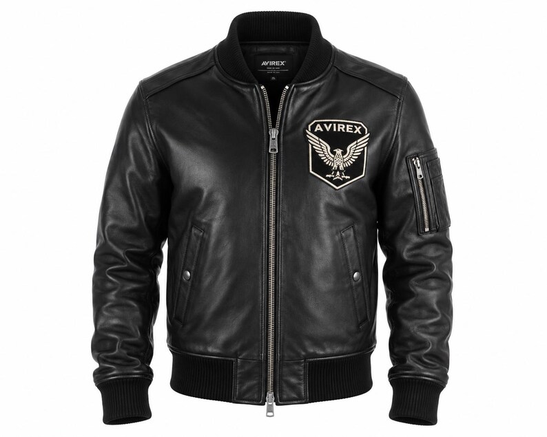 May include: A black leather bomber jacket featuring a ribbed collar, cuffs, and hem. The front includes a central silver zipper, two side pockets with snap closures, and a patch on the left chest displaying an eagle emblem and the text AVIREX.