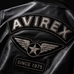 May include: A black leather jacket featuring a large arched patch on the back with the text AVIREX in silver lettering. Below this, a winged star emblem is centered above a rectangular patch displaying the text SINCE 1975 in silver font.