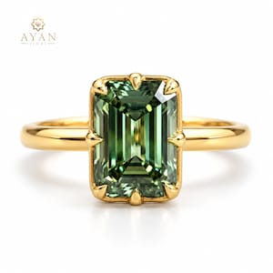 Op de afbeelding: Een gouden ring met een grote, rechthoekige, smaragdgeslepen groene edelsteen in een klauwzetting. De ringband is van glad, gepolijst goud. Het logo AYAN JEWELS is zichtbaar in de linkerbovenhoek van de afbeelding, boven de ring.