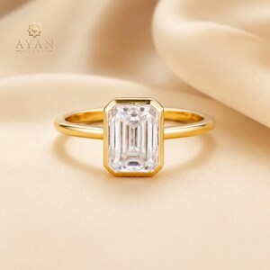 May include: A gold ring featuring a clear, rectangular emerald-cut gemstone set in a smooth bezel setting. The band is polished gold. The text AYAN JEWEL is visible in the upper left corner, accompanied by a small star-shaped logo icon.