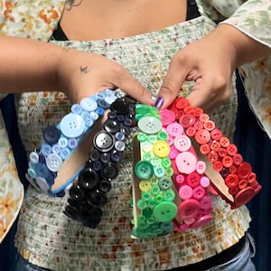 May include: Four headbands decorated with various sizes of round buttons in distinct color palettes. The headbands feature blue, black, green, and red-to-pink gradients, with each button securely attached to the surface of the headband frame.