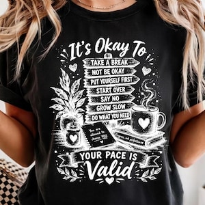 以下が含まれることがあります： It's Okay To, Take A Break, Not Be Okay, Put Yourself First, Start Over, Say No, Grow Slow, Do What You Need, Your Pace Is Validというテキストが書かれた木製の看板の白いグラフィックプリントが施された黒いTシャツ。
