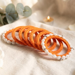 May include: A set of circular bangles featuring a vibrant orange yarn-wrapped base. Each bangle is accented with a series of small, round, white faux pearls spaced evenly around the circumference, secured by tiny silver-toned metal beads.
