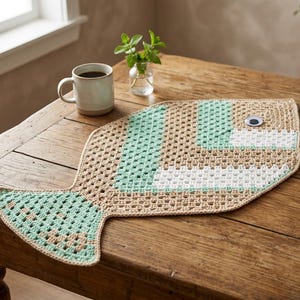 Könnte beinhalten: Eine fischförmige Häkelmatte mit einem geometrischen Muster aus beigem, mintgrünem und weißem Garn. Die Matte verfügt über ein einzelnes, kreisförmiges Wackelauge im Kopfbereich und zeigt eine detaillierte Konstruktion mit offenen Maschen.