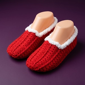 May include: A pair of bright red crochet slippers featuring a textured ribbed pattern and a soft white fuzzy trim around the ankle opening. The slippers are displayed on beige mannequin foot forms, showcasing the deep red yarn and white cuff.