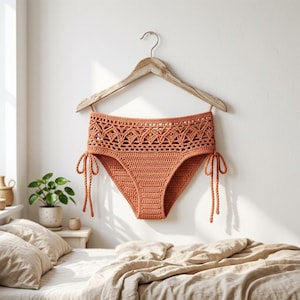 May include: A high-waisted terracotta-colored crochet bikini bottom featuring an intricate geometric pattern along the waistband and adjustable side ties with dangling cords. The garment is displayed on a wooden hanger against a plain wall.