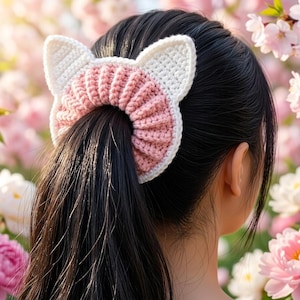 Könnte beinhalten: Ein gehäkeltes Haargummi mit einer gerippten rosa Mitte und weißen Akzenten in Form von Katzenohren. Das Accessoire ist um einen Pferdeschwanz aus dunklem Haar gebunden und zeigt eine strukturierte Garnkonstruktion mit deutlichen Farbblöcken.
