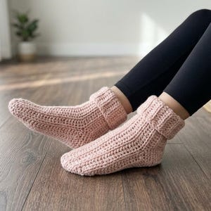 May include: A pair of light pink, hand-crocheted ankle socks featuring a thick, ribbed texture and a folded cuff design. The socks are crafted with a visible yarn stitch pattern and provide a snug fit around the feet, extending just above the ankle.