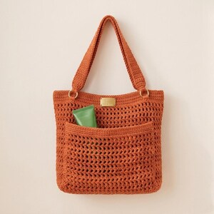 Pode incluir: Uma bolsa tote de crochê em laranja queimado com padrão de tecido aberto, duas alças resistentes presas com anéis de metal dourado e uma placa retangular de metal dourado centralizada no topo. Um tubo verde está inserido no bolso frontal.