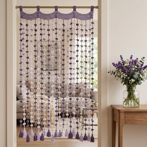 Pode incluir: Uma cortina de porta em crochê com uma borda superior festonada e múltiplos fios verticais adornados com pequenos nós florais uniformemente espaçados em tons de branco, roxo claro e roxo escuro, finalizados com borlas na base.
