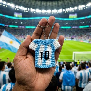 Può includere: Un piccolo portachiavi all'uncinetto a forma di maglia sportiva con strisce verticali azzurre e bianche. La maglia presenta il numero 10 ricamato in nero sul davanti ed è fissata a un anello di metallo argentato tramite una corta catenella.