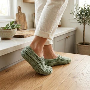 May include: A pair of sage green crochet slippers featuring a textured, ribbed stitch pattern. The slippers have a low-cut design that covers the foot up to the ankle, with a rounded toe and a thick, hand-stitched construction in a soft yarn.