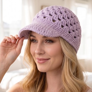 Puede incluir: Una gorra de béisbol de ganchillo en color lavanda claro que presenta un patrón de tejido abierto en toda la copa y una visera sólida de punto apretado. El gorro está confeccionado con hilo de textura visible y un color pastel uniforme.
