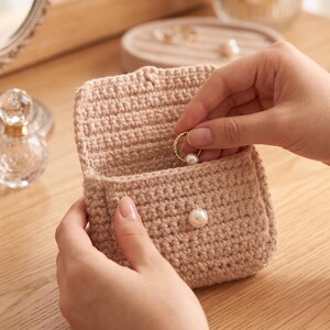 May include: A small, beige, crocheted pouch with a flap closure secured by a single white pearl button. The pouch is constructed from a textured yarn and is designed to hold small items like jewelry, such as the gold ring with a pearl being placed inside.