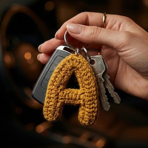 Puede incluir: Un llavero con la letra A tejida a ganchillo en color amarillo mostaza, unido a una anilla de metal plateado. El llavero también incluye un mando de coche de plástico negro y dos llaves de metal plateado. La letra de hilo tiene textura.