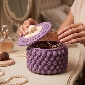 Può includere: Un portagioie rotondo all'uncinetto di colore viola con un motivo a punto nocciolina testurizzato sui lati. Il coperchio è viola con un pomello centrale color crema. All'interno della scatola ci sono diversi braccialetti sottili in metallo dorato.
