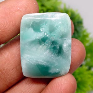 May include: A rectangular polished gemstone featuring a smooth surface with a marbled appearance in shades of teal, mint green, and white. The stone displays translucent areas and internal crystalline textures throughout its polished face.