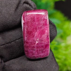May include: A rectangular, polished gemstone with a deep magenta and ruby red color. The stone features a smooth, slightly rounded surface with visible internal crystalline textures and natural inclusions throughout its vibrant, saturated body.
