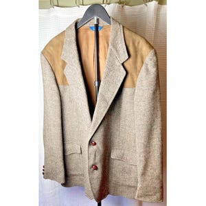 May include: A beige and brown herringbone blazer featuring tan suede shoulder patches, a notched lapel, two front flap pockets, and two dark brown buttons. The interior is lined with a solid tan fabric and includes a blue brand label at the neck.
