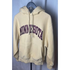 Puede incluir: Una sudadera con capucha de color amarillo pálido que presenta la palabra MINNESOTA impresa en una fuente arqueada de color granate oscuro en el pecho. La prenda incluye un bolsillo canguro frontal, capucha con cordones blancos y mangas largas.