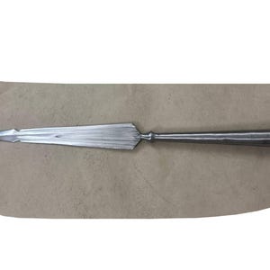 Op de afbeelding: Een zilverkleurige metalen speerpunt met een scherpe, puntige top en een lang, taps toelopend blad met een centrale richel. De basis van het blad is verbonden met een cilindrische, holle huls die bedoeld is voor bevestiging aan een houten schacht.