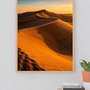 May include: A framed vertical wall art print featuring a desert landscape with golden sand dunes under a sunset sky. The image displays a sharp ridge line of sand casting long shadows, with a gradient of orange, yellow, and blue hues in the sky.