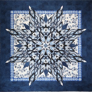 May include: A square quilt featuring a large central eight-pointed star in shades of navy, light blue, and white. The star is set against a floral patterned background, framed by a border of small blue triangles and a solid navy blue outer edge.