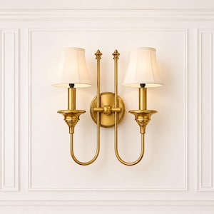 May include: A double-arm wall light featuring a polished brass finish, two vertical rods with finial tops, and two off-white tapered fabric shades. The fixture is mounted on a circular brass backplate with a central sphere detail.