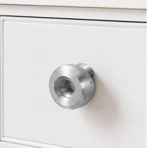 Puede incluir: Un pomo de mueble circular de metal plateado cepillado con un centro hundido, montado en un frente de cajón blanco liso. El pomo presenta un borde exterior biselado y una base cilíndrica, aportando un acento metálico a la superficie blanca.