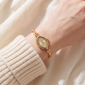 May include: A gold-tone quartz wristwatch featuring an elongated marquise-shaped case with a champagne dial and the brand name Lucx printed in black. The watch band consists of delicate, interconnected floral-shaped metal links in gold.