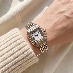 May include: A two-tone wristwatch with a square silver-tone case and a gold-tone bezel. The white dial features black Roman numeral hour markers, blue hands, and the text GREALY QUARTZ. The bracelet consists of alternating silver and gold links.