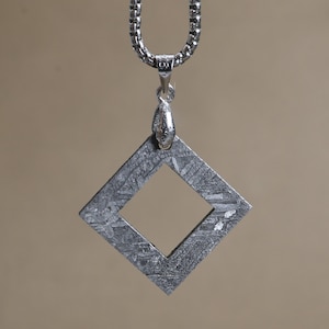 May include: A square-shaped pendant with a textured, metallic grey surface featuring a cross-hatched pattern, suspended from a silver-toned box chain. The pendant has a central square cutout and is attached to the chain by a small silver bail.
