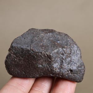 Puede incluir: Una roca de color marrón oscuro de forma irregular con una superficie rugosa, picada y texturizada. El objeto presenta un aspecto denso y metálico con pequeñas hendiduras y un acabado mate, mostrando variaciones naturales en su color.