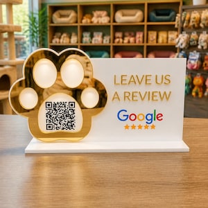 May include: A white rectangular sign featuring a gold-colored paw print cutout with a QR code in the center. The sign displays the text LEAVE US A REVIEW above the Google logo and five gold stars, designed for customer feedback.