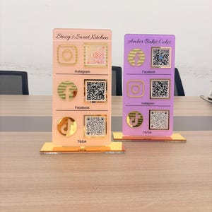 May include: Two rectangular acrylic social media signs with gold stands. The peach sign reads Stacy's Sweet Kitchen, and the purple sign reads Amber Bakes Cakes. Both feature gold social media icons and QR codes for Instagram, Facebook, and TikTok.