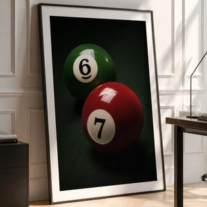 May include: A framed wall art print featuring a dark green billiard ball with the number six and a red billiard ball with the number seven. The balls are positioned against a dark, textured background, creating a high-contrast composition.