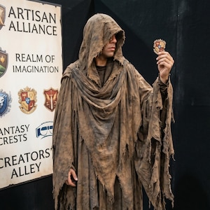 May include: A floor-length, distressed brown hooded robe with a tattered, uneven hemline and frayed edges. The garment features a draped cowl neckline and long, wide sleeves. The fabric has a weathered, textured appearance with dark shading.