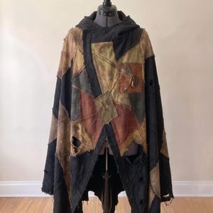 Op de afbeelding: Een cape met capuchon gemaakt van een patchwork van zwarte, bruine en olijfgroene stoffen panelen. Het kledingstuk heeft zichtbare gerafelde randen, versleten naden en verschillende kleine gaatjes in het materiaal, wat zorgt voor een verweerde uitstraling.