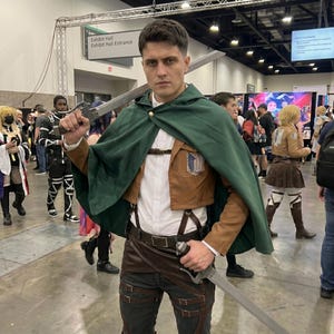 May include: A person wearing a Survey Corps cosplay outfit featuring a dark green hooded cape, a cropped tan jacket with a blue and white wing emblem, a white button-down shirt, dark trousers, and brown leather harness straps, holding two swords.