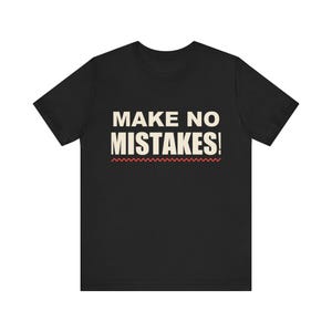 May include: A black short-sleeved t-shirt featuring the text "MAKE NO MISTAKES!" printed in large, bold, off-white capital letters across the chest, with a thin, horizontal, wavy red line positioned directly underneath the text.