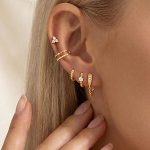 Peut inclure: Un ensemble de quatre boucles d'oreilles dorées portées sur une seule oreille. La collection comprend un petit clou à trois pierres, une manchette à double anneau, un anneau fin avec une pierre et une petite créole avec une fine chaînette.